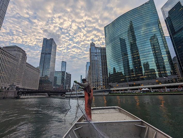 A person in dirty orange overalls stands at the bow of a flat-bottom boat holding a portion of netting above the water.